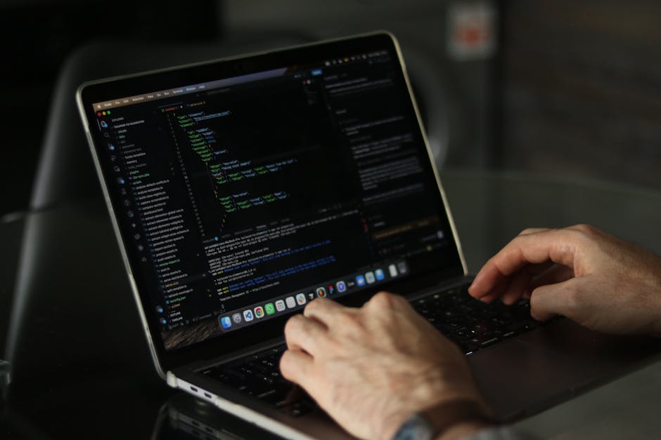 Close-up view of a person's hands working on a MacBook laptop placed on a dark table, displaying lines of code in a programming environment with a black background and colorful syntax highlighting. Th