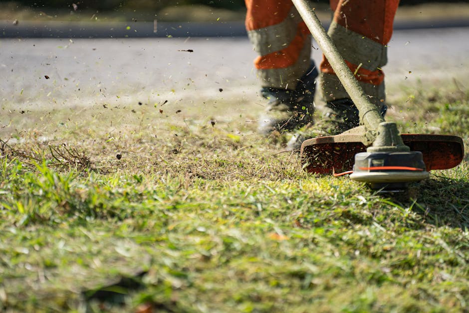 A person wearing orange work trousers and dark shoes is using a petrol-powered grass trimmer to clear overgrown grass and weeds in a garden. The garden features a mix of trimmed and untrimmed grass, w