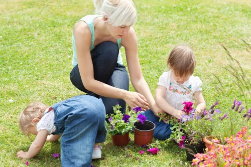 A woman with short blond hair, dressed in a light blue sleeveless top and dark jeans, is kneeling on a well-maintained grassy lawn, engaged in planting or tending to garden flowers alongside two young children. One child, with short blond hair and wearing a white shirt with a floral pattern and blue jeans, is seated on the grass, carefully examining or handling vibrant purple and pink flowers. The other child, also with light hair and dressed in a white top with a different floral pattern, is lying on the grass, leaning forward, and appears to be reaching towards the flowers. Around them are several small pots and containers, some filled with soil and plants, indicating active gardening. The garden area features a lush, green lawn with healthy, dense grass, bordered by flower beds with bright, colorful blooms. In the background, there are some bushes or small trees, suggesting a landscaped outdoor space in a residential garden setting, typical of Thamesmead. The weather appears bright and sunny, creating a warm outdoor environment suitable for gardening activities.